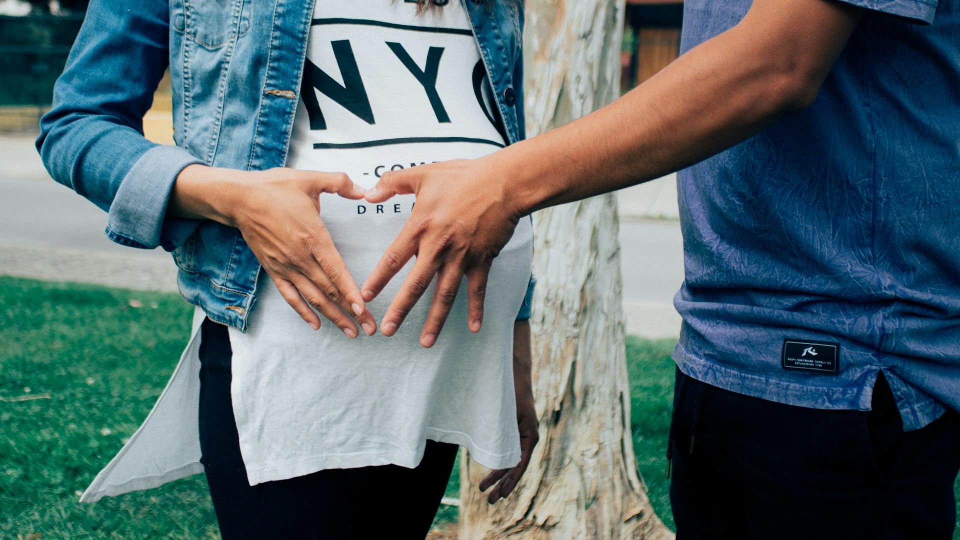 Expectant parents forming heart on belly, celebrating pregnancy outdoors.