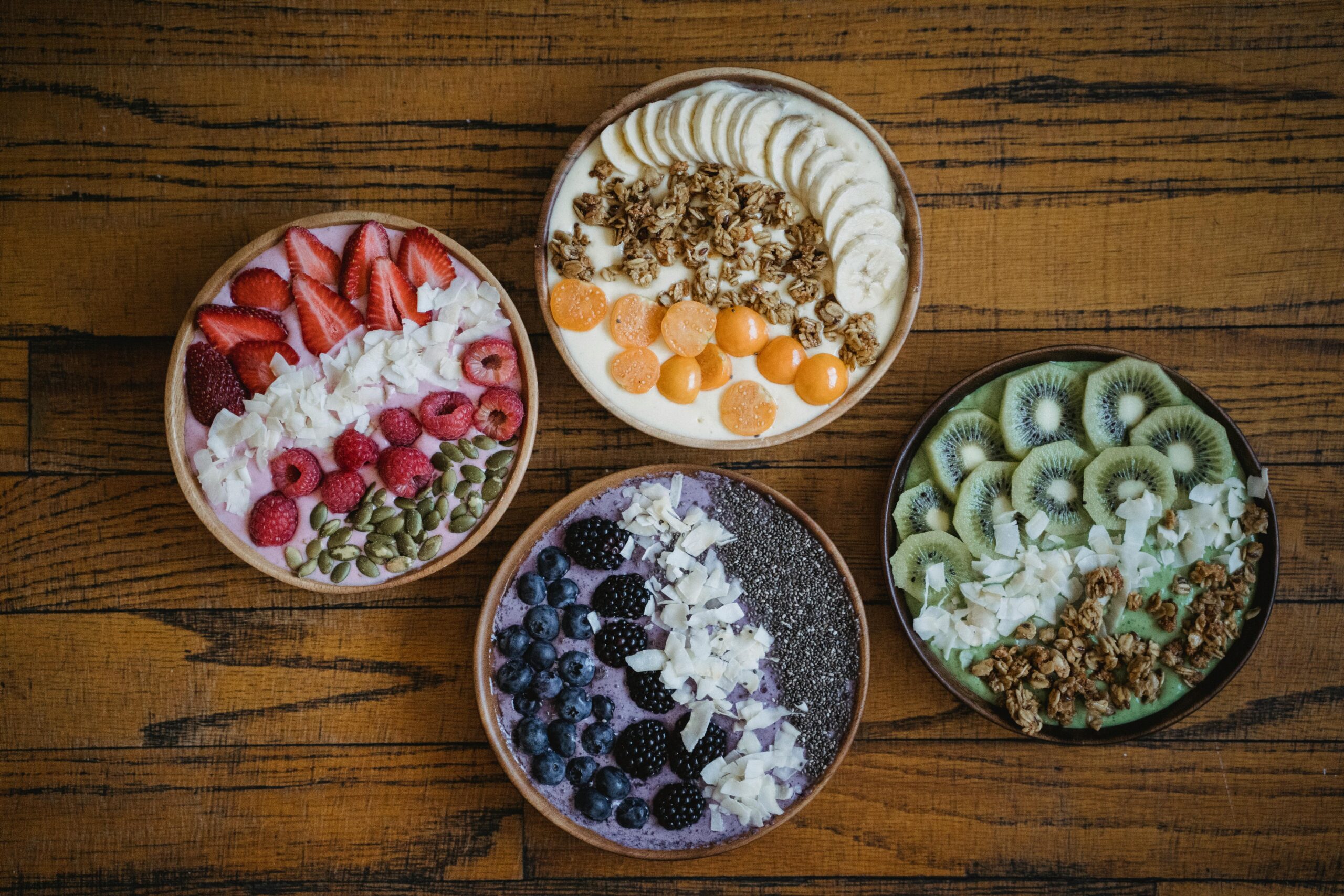 Four vibrant smoothie bowls with fruits, nuts, and granola on a wooden table.